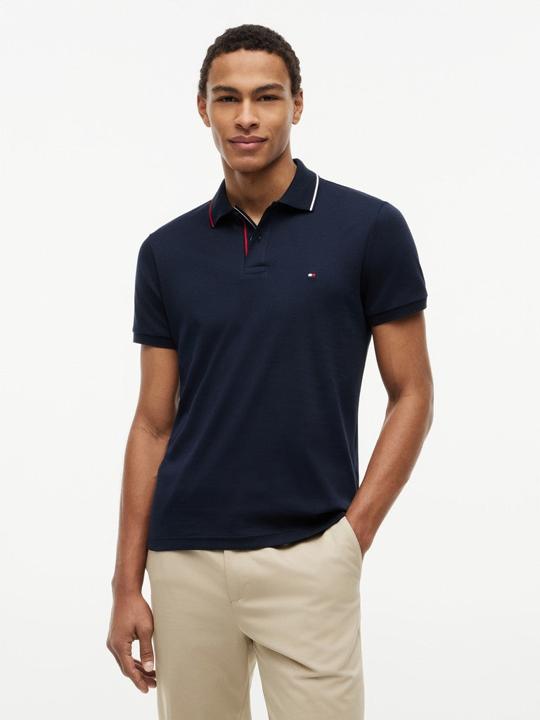 A man wearing a navy blue short-sleeve polo shirt with a white and red striped collar, paired with beige trousers, photographed against a plain white studio background.