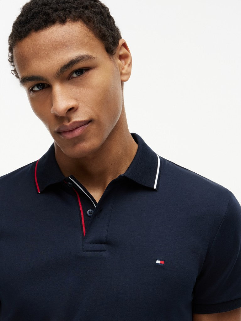 A close-up of a man wearing a navy blue Tommy Hilfiger polo shirt with white and red striped trim on the collar and a small embroidered flag logo on the chest.