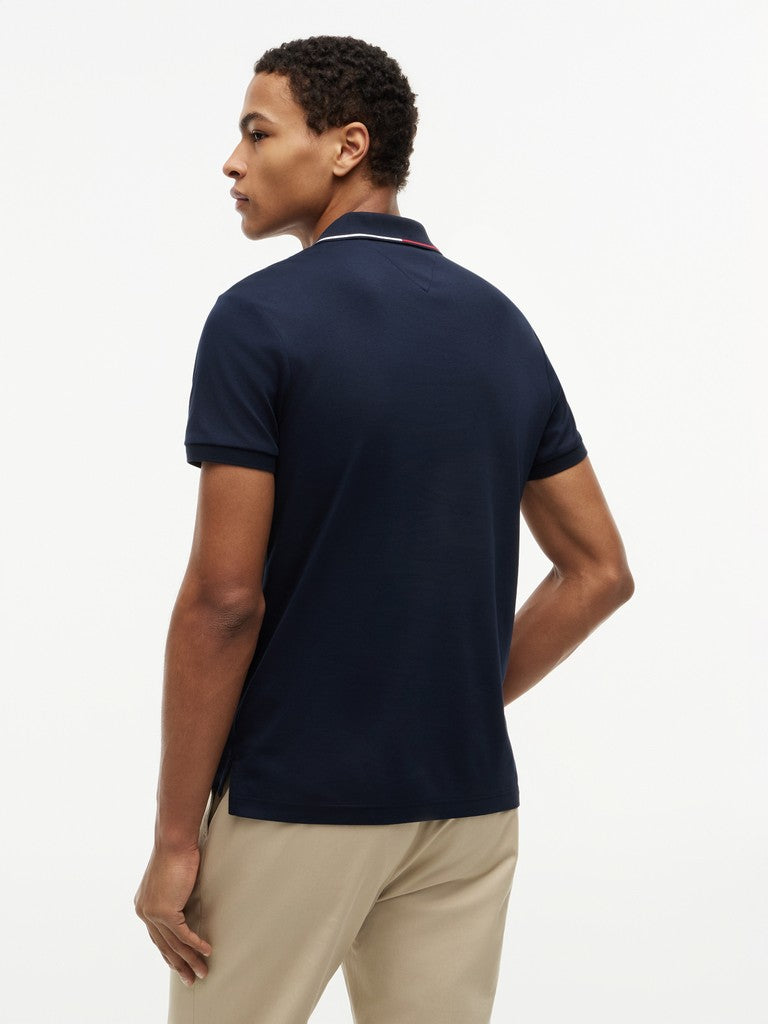 A man wearing a navy blue short-sleeve polo shirt with a thin red and white stripe detail on the collar, paired with beige trousers, shown from a three-quarters back view against a plain white background.