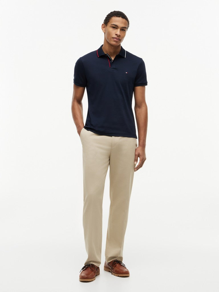 A man modeling a navy blue short-sleeve polo shirt with a red and white striped collar, paired with beige trousers and brown leather shoes, against a plain white studio background.