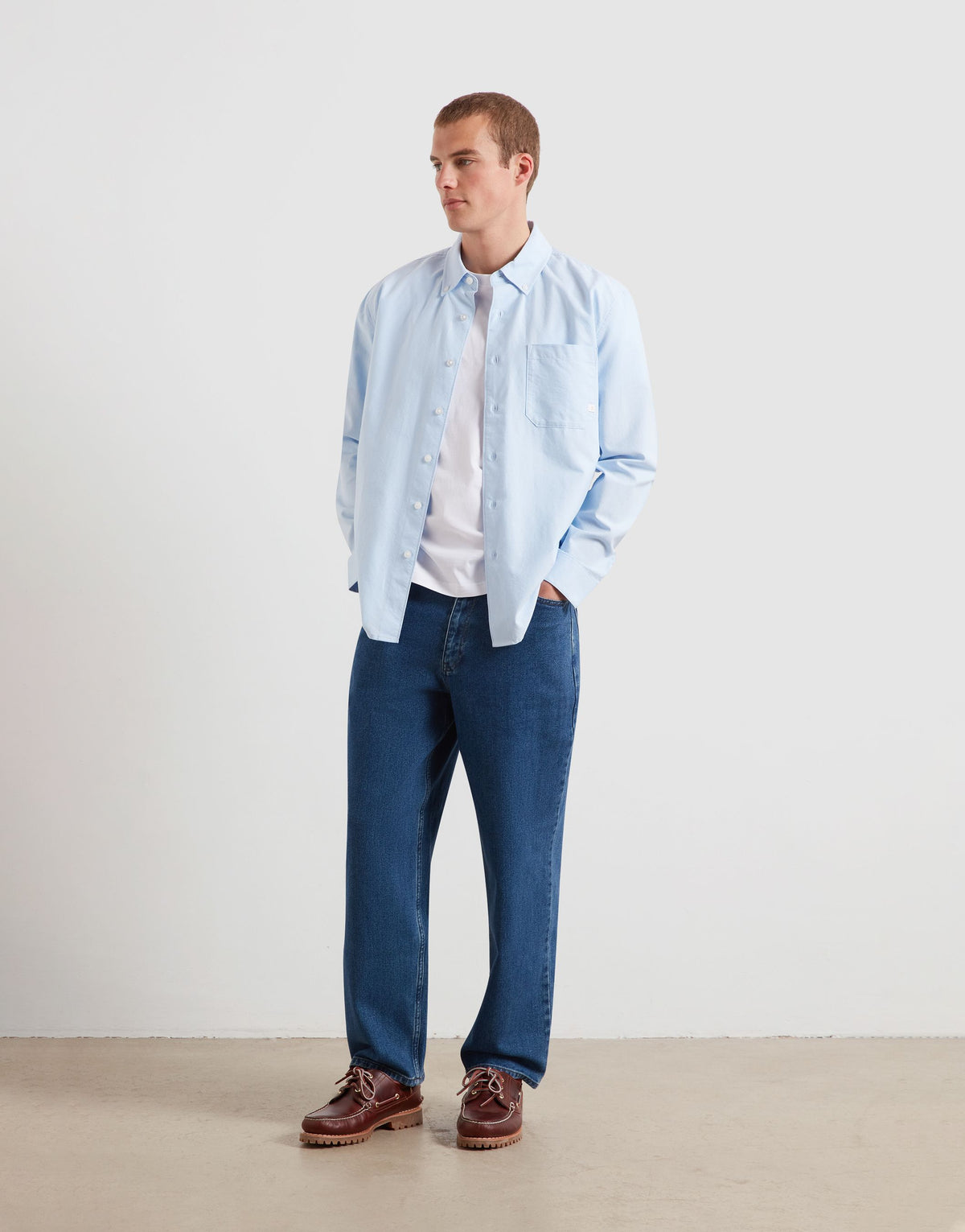 A man wearing a light blue button-down shirt over a white t-shirt, paired with dark blue jeans and brown leather shoes, standing against a plain off-white studio background.