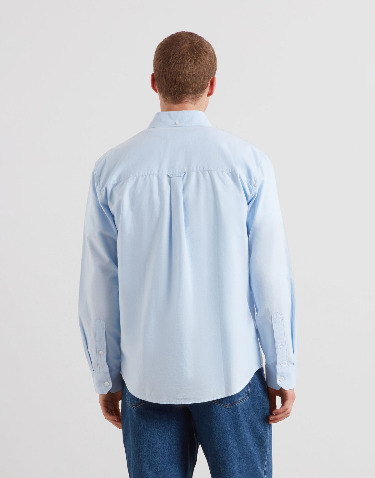 A man viewed from behind wearing a light blue long-sleeve button-up shirt and blue jeans, standing against a plain light gray studio background.