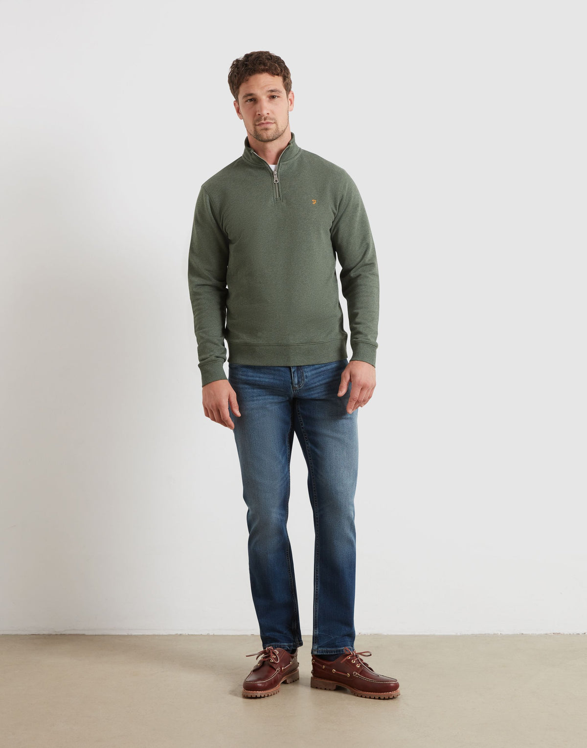 A man wearing an olive green quarter-zip sweater, blue jeans, and brown leather shoes, standing against a plain light gray studio wall.