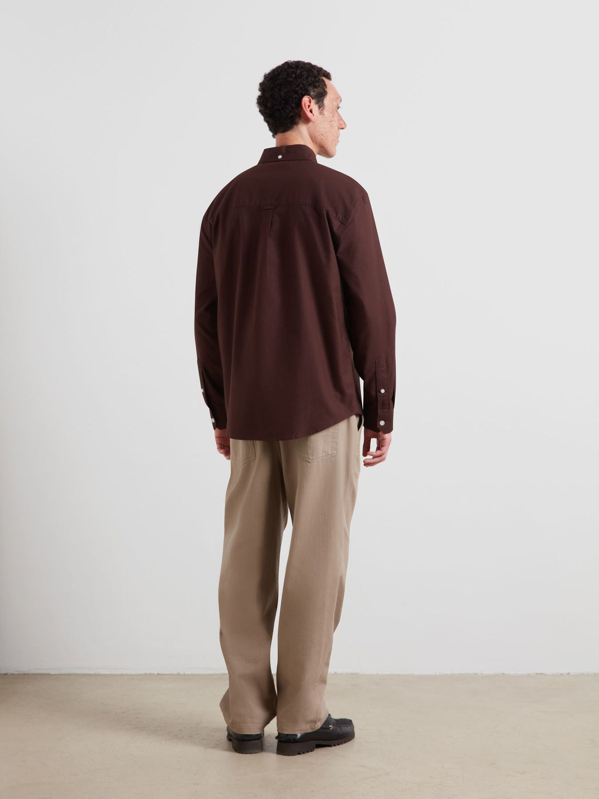 A man is shown from the back wearing a dark brown long-sleeve button-up shirt and beige trousers, standing against a plain light gray studio wall.