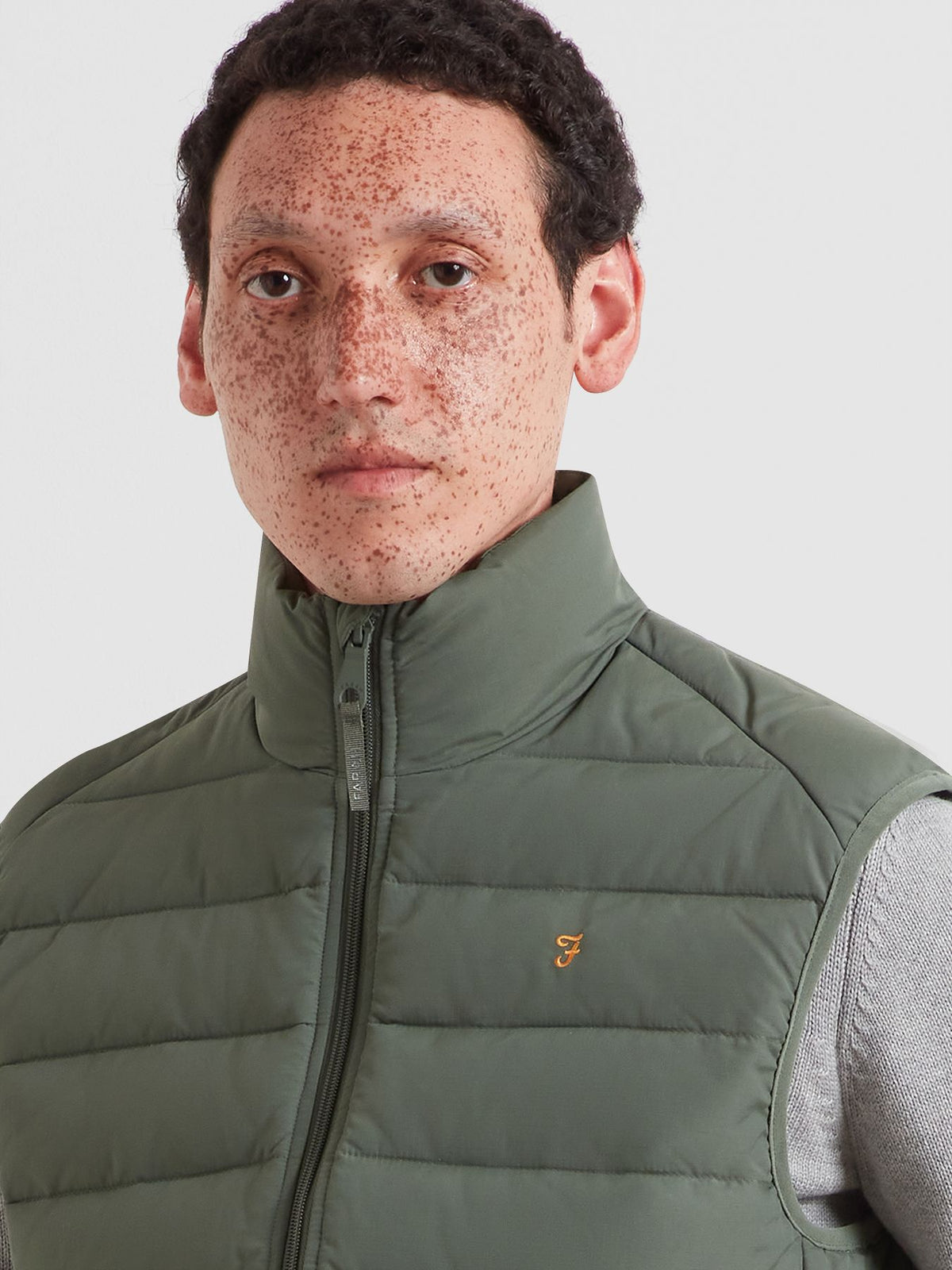 A close-up of a man wearing an olive green puffer vest over a gray knit sweater, against a light gray background.