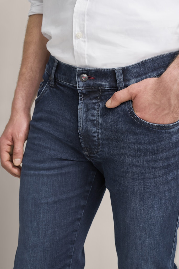 A close-up of a person wearing dark blue slim-fit jeans with a white button-up shirt tucked in, showing the front pocket and hand in the pocket.