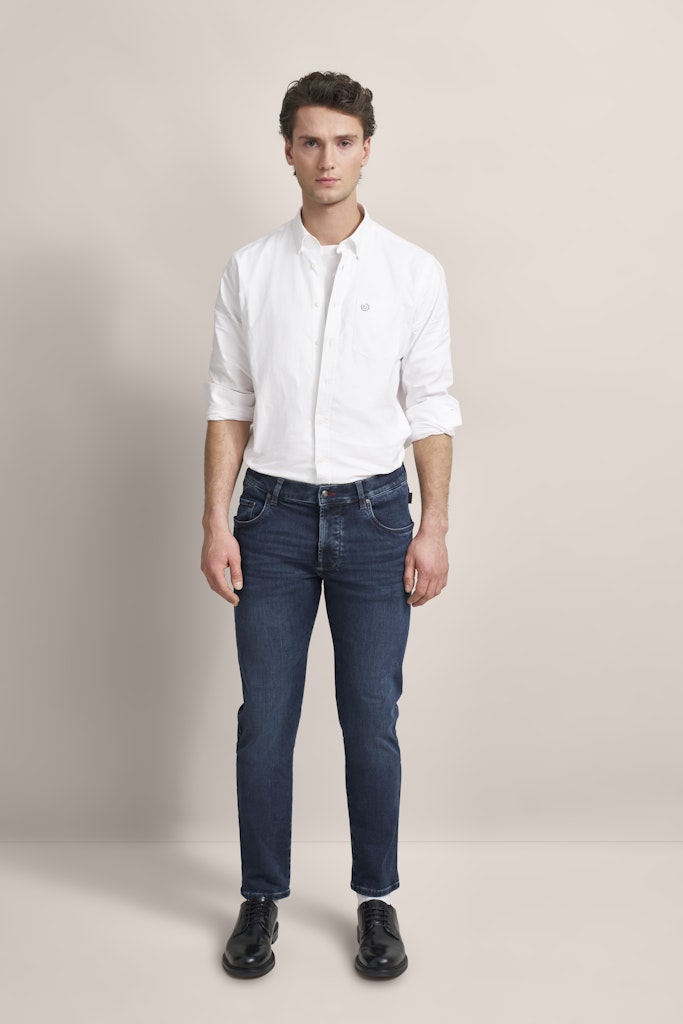 A man modeling a white long-sleeve button-down shirt and dark blue slim-fit jeans with black shoes, standing against a neutral beige studio background.