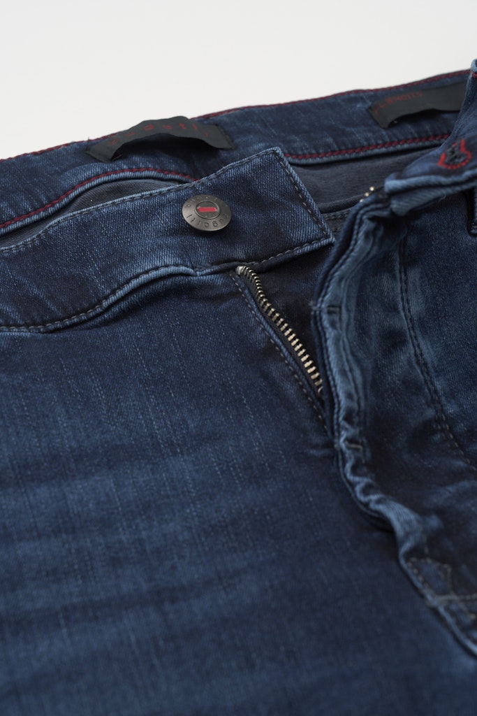 Close-up of dark blue denim jeans showing the waistband, button closure, zipper, and red contrast stitching against a light background.
