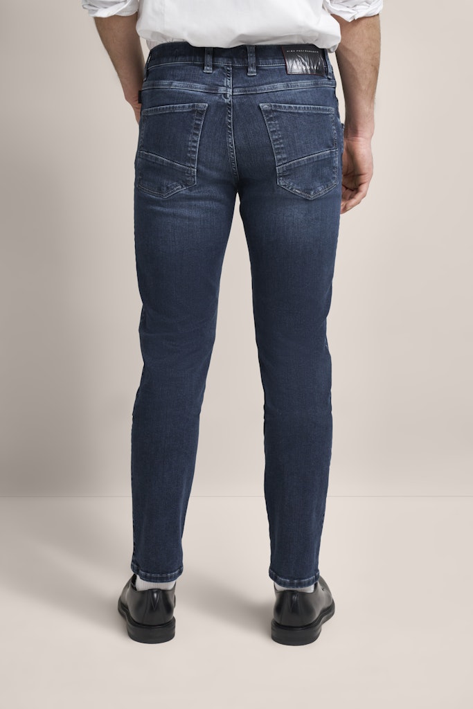 Rear view of a person wearing dark blue slim-fit jeans with a white shirt and black shoes, standing against a light beige studio background.