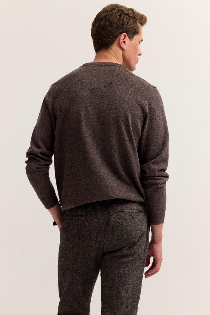 A man viewed from the back wearing a dark brown long-sleeve knit sweater and dark charcoal gray trousers, standing against a plain off-white studio background.