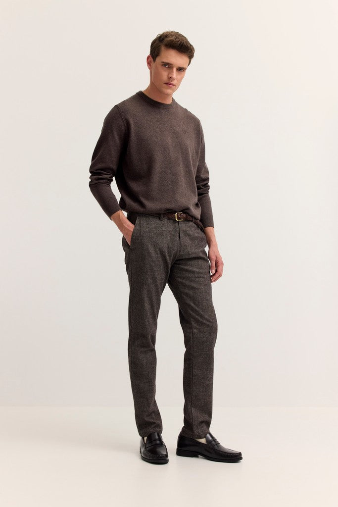 A man modeling a dark brown crewneck sweater and dark gray textured trousers with black leather shoes, standing against a plain light gray studio background.