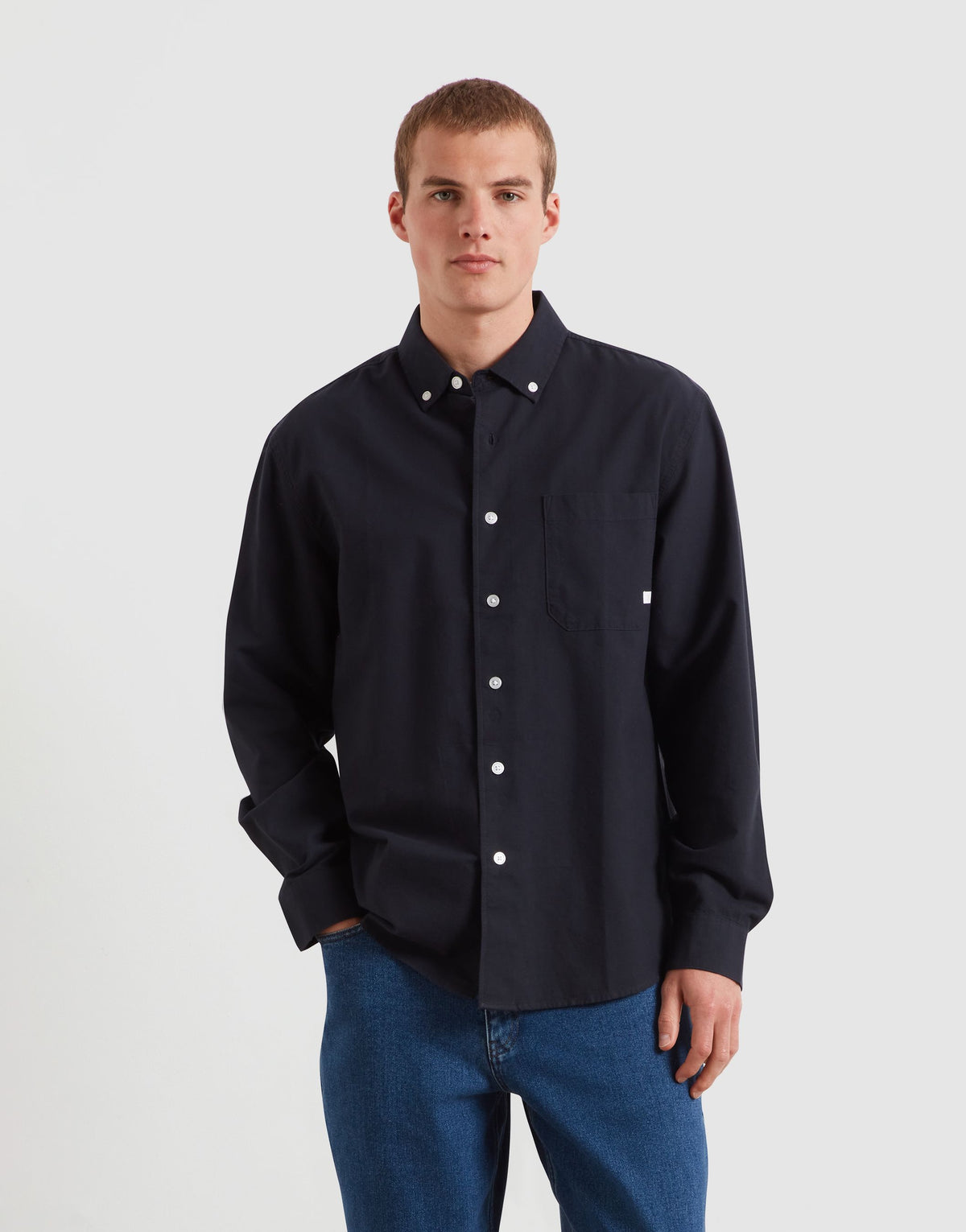 A man wearing a dark navy blue long-sleeve button-down shirt with white buttons and blue jeans, standing against a light gray studio background.