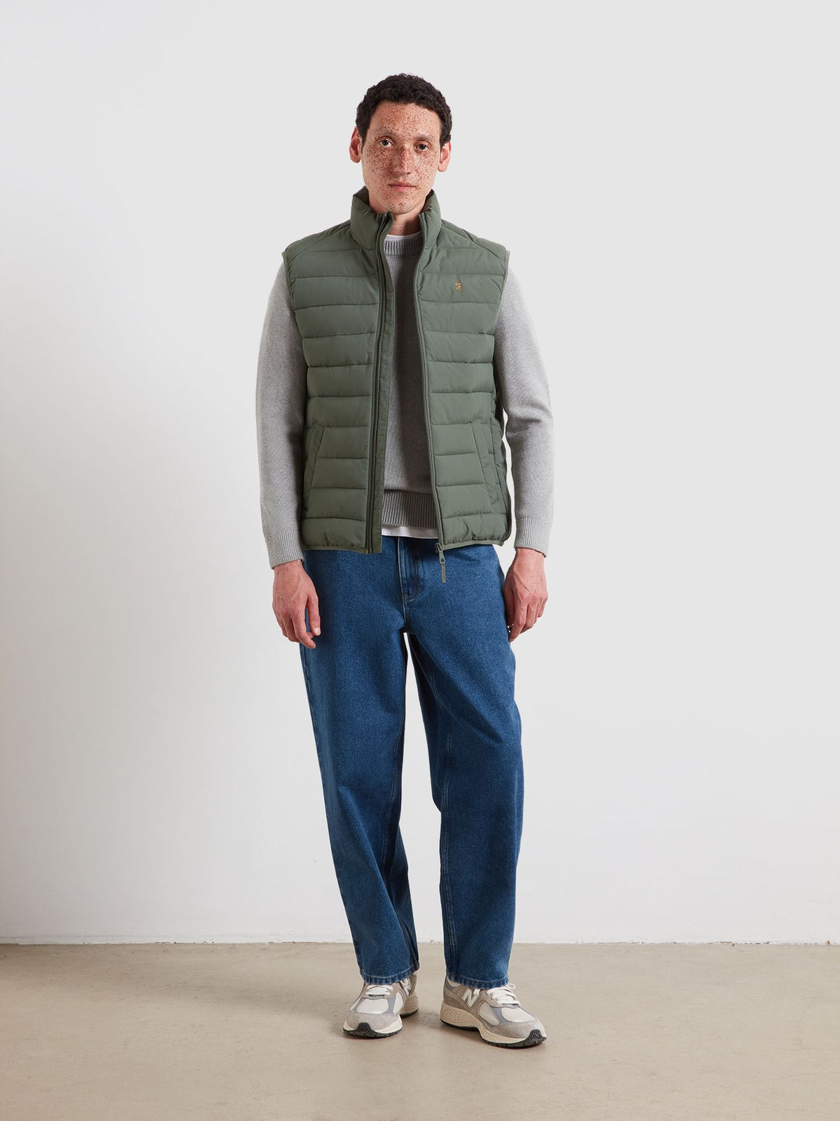 A man wearing a sage green puffer vest over a gray long-sleeve shirt, blue jeans, and white sneakers, standing against a plain off-white wall.