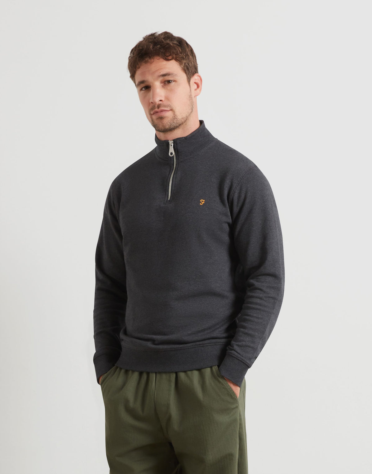 A man wearing a dark charcoal gray quarter-zip sweater with a small gold F embroidered logo on the chest, paired with olive green pants, against a light gray studio background.