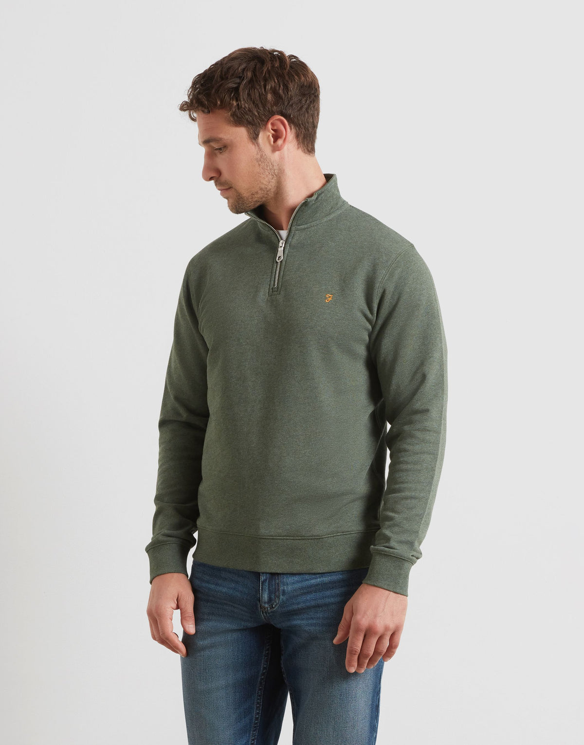 A man wearing an olive green quarter-zip pullover sweater with a small embroidered logo on the chest, paired with blue jeans, photographed against a light gray studio background.