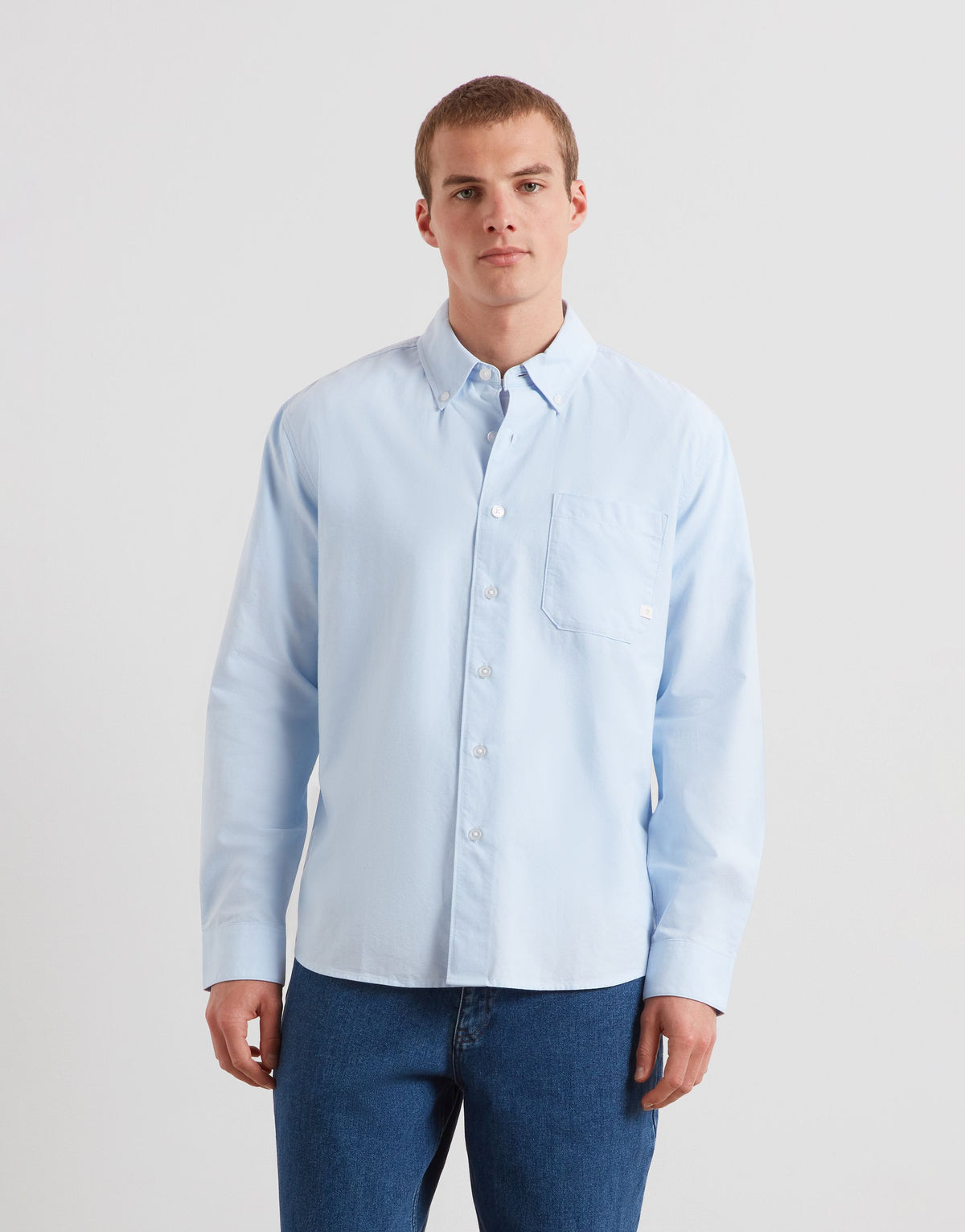 A man wearing a light blue long-sleeve button-down shirt and blue jeans, standing against a plain light gray studio background.