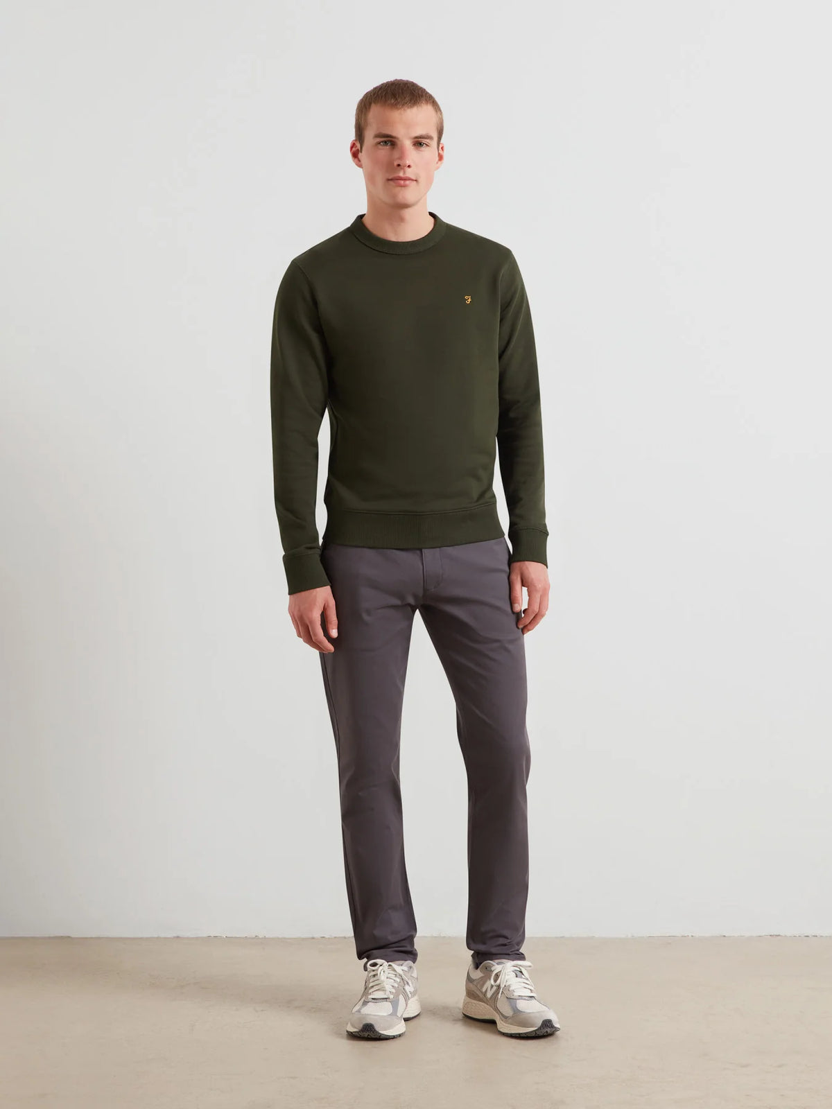 A man wearing an olive green crewneck sweater, dark gray trousers, and white sneakers, standing against a plain light gray wall.