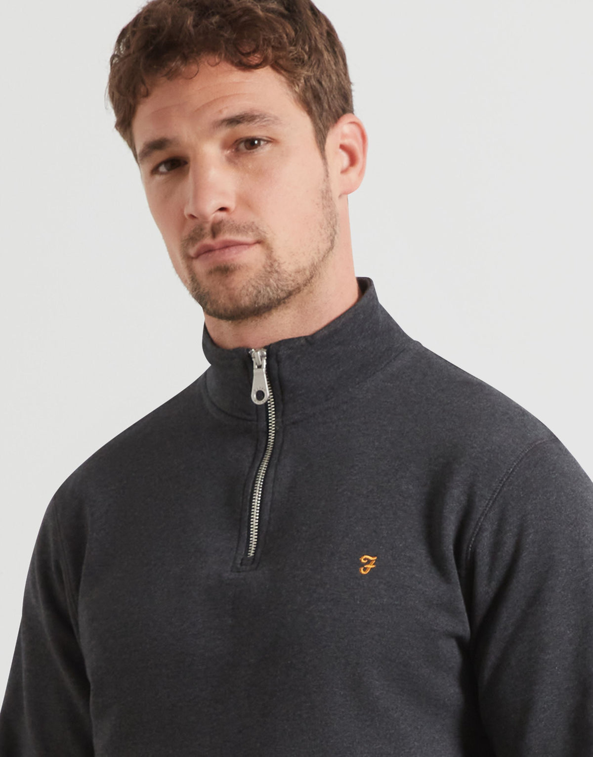 A man wearing a dark charcoal gray quarter-zip pullover sweater with a small gold F embroidered logo on the left chest, photographed against a light gray studio background.