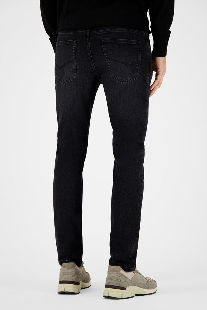 Rear view of a person wearing slim-fit black jeans with a black long-sleeve top and beige sneakers, photographed against a clean light gray studio background.