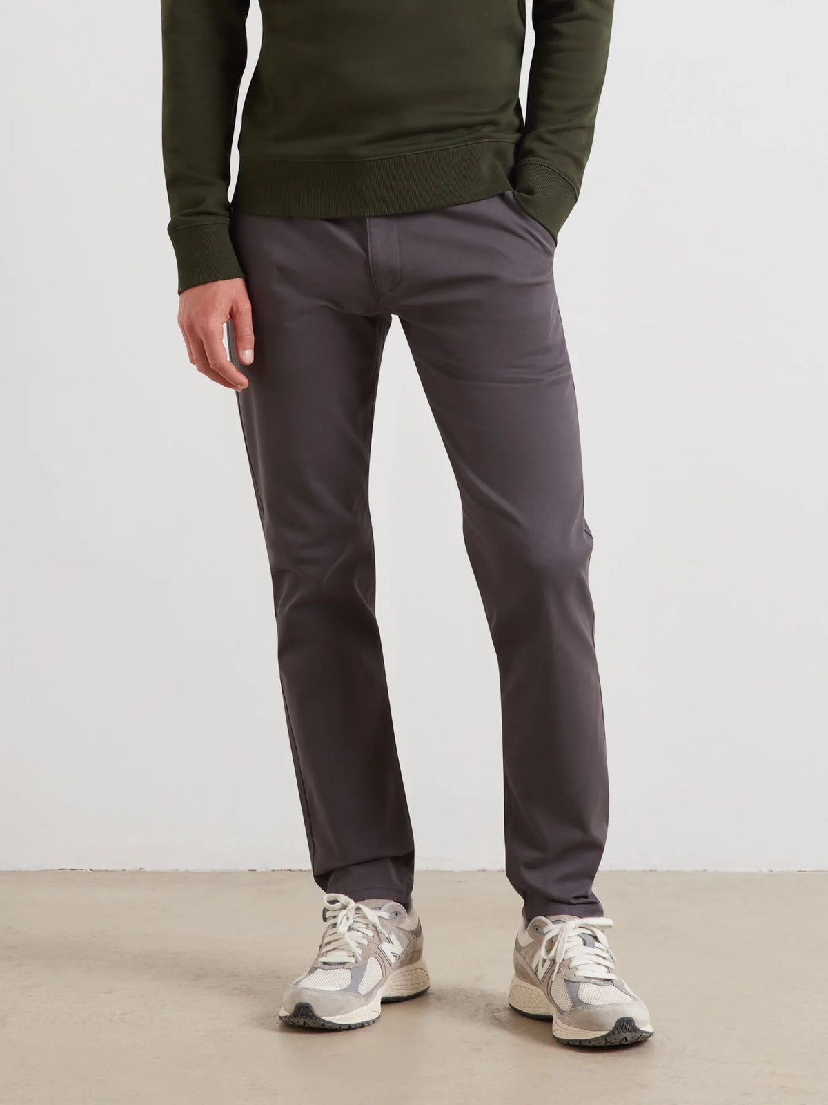 A person wearing slim-fit dark gray trousers, an olive green long-sleeve top, and light gray sneakers, standing against a plain light gray wall.