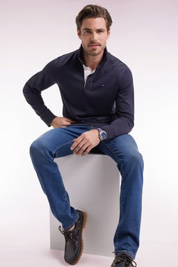Man sitting on a white block wearing a navy rugby top, blue jeans, and black shoes.