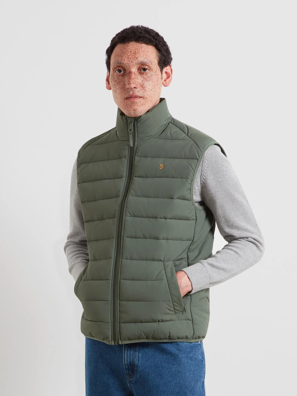 A man wearing an olive green puffer vest over a light gray long-sleeve shirt and blue jeans, standing against a light gray studio background.