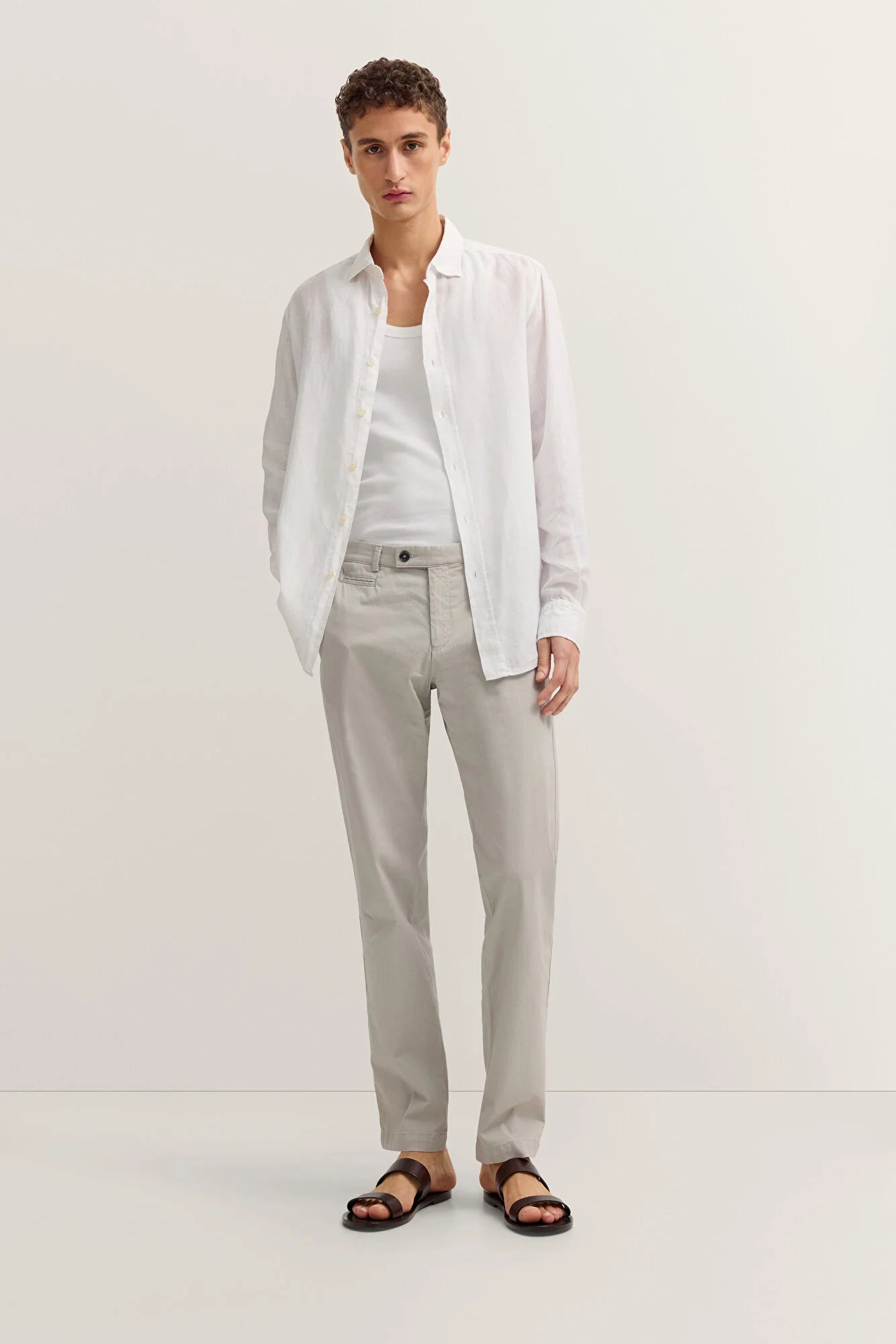 Man wearing a white shirt open with a white T-shirt, light grey chinos and brown sandals against a light grey background